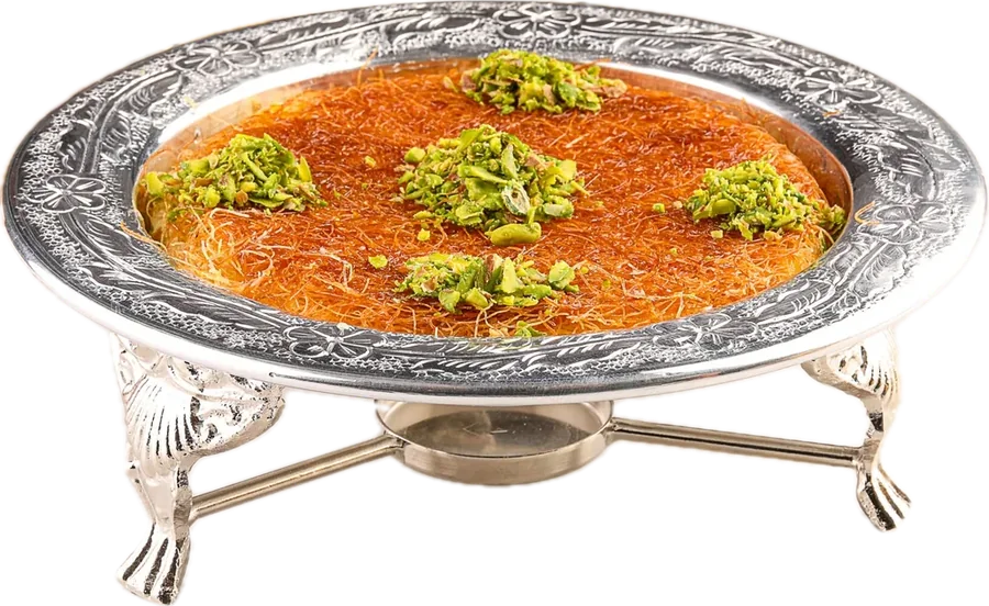 A round knafeh on a footed silver stand, topped with clusters of green pistachio.